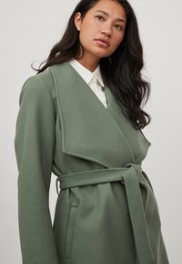 Abrigo verde de mezcla de lana con cuello chal, cinturón de lazo y bolsillos laterales. Textura suave con una silueta ajustada, llevado sobre una camisa blanca con cuello.