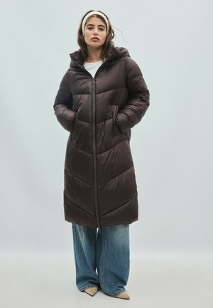 Femme portant un long manteau matelassé marron foncé avec les mains dans les poches, un jean large bleu, des chaussures beiges et un bandeau blanc sur fond gris.