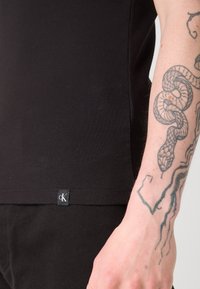 Črn rebrast top z majhnim CK logotipom pri robu, v kombinaciji s črnimi hlačami. Roka je vidna, na njej pa je podrobna tetovaža kače.