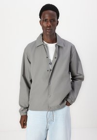 Grå lättviktsjacka med knäppt framtill, mandarin-krage, elastiska ärmslut och dragsko i nederkant, matchad med en vit T-shirt och ljusa jeans.