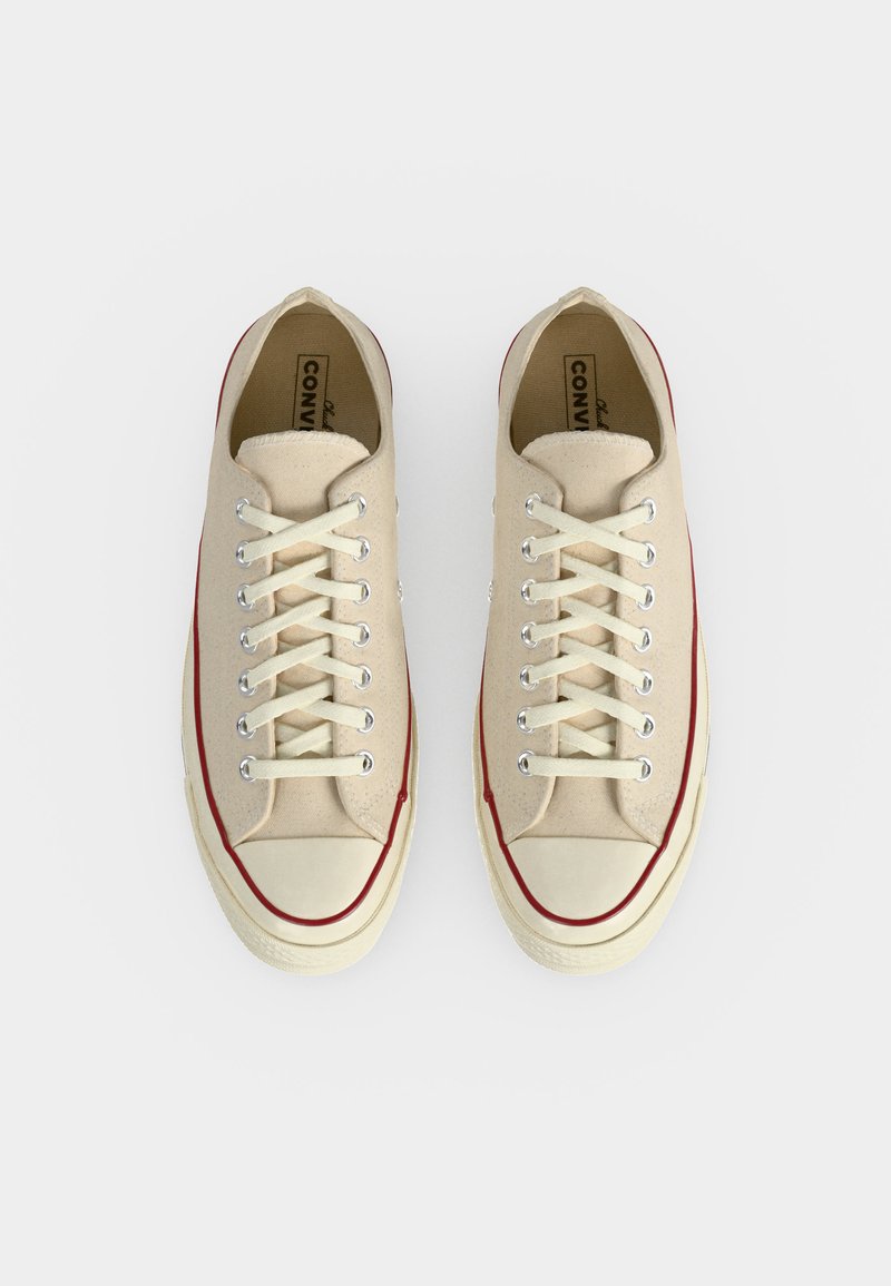 Coppia di sneakers basse in tela color crema con lacci bianchi e striscia rossa lungo la suola, vista dall'alto su sfondo bianco.