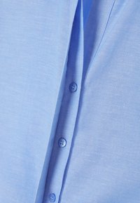 Světle modrá košile na knoflíky s jemnou, texturovanou látkou a šesti sladěnými knoflíky podél légy.