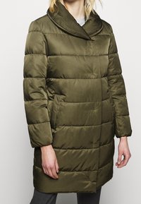 Olivengrøn polstret jakke med høj krave, quiltet tekstur, sidelommer og knælangt design. Har en frontknaplukning.