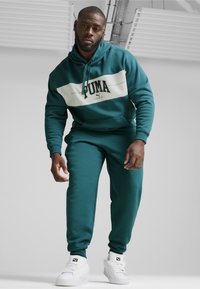 Felpa teal con striscia bianca che presenta il logo "PUMA", abbinata a pantaloni sportivi teal coordinati e sneakers bianche. Tessuto morbido, vestibilità comoda.