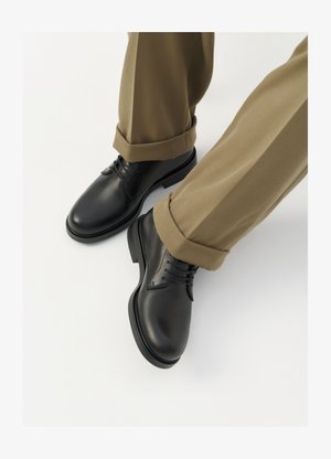 Scarpe da uomo in pelle nera con una finitura liscia, dotate di lacci e punta arrotondata, abbinate a pantaloni khaki arrotolati.