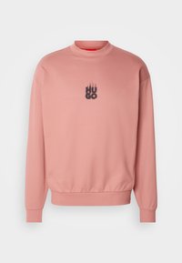 Rózsaszín pulóver magas nyakkal és hosszú ujjakkal. Kontrasztos fekete "HUGO" logó található egy láng design fölött. Sima textúra.