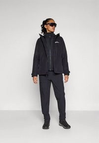 Fekete kültéri dzseki kapucnival, fekete zipzáras felsőréteg fölött viselve, fekete nadrággal és masszív fekete cipővel párosítva. Mellrészen logóval.