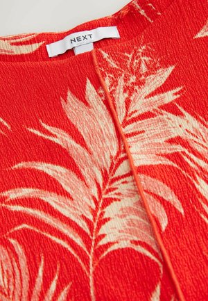 Červená textúrovaná tkanina s krémovými vzormi listov, so stredovým švom a štítkom označeným "NEXT" na výstrihu.