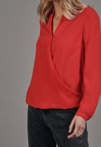 Blusa rossa a maniche lunghe con scollatura a V e design avvolgente. Realizzata in un tessuto liscio con dettaglio a bottoni all'ascella.
