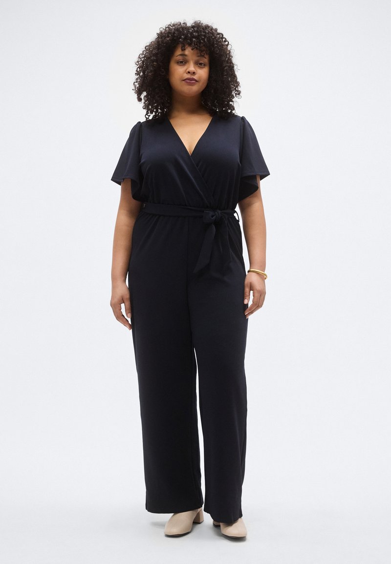 Tengerészkék, átlapolt stílusú overál rövid, fodros ujjakkal, öves derékkal és bőszárú nadrággal. Sima anyag, puha tapintású.