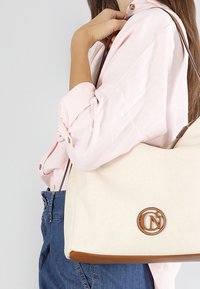 Bolso de lona beige con detalles en cuero marrón, que presenta un logo circular. El bolso tiene una forma estructurada y una correa de hombro ajustable.