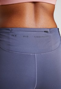 Zbliżenie na dolną część pleców i talię osoby noszącej fioletowe legginsy z drobnym nadrukiem oraz zamkniętą kieszonką na zamek błyskawiczny przy pasie.