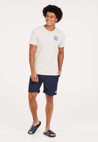 Beige t-shirt met korte mouwen en een blauwe graphic op de borst, gecombineerd met donkerblauwe shorts en zwarte slippers. Eenvoudig ontwerp en casual pasvorm.