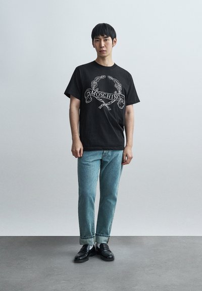 Μαύρο βαμβακερό μπλουζάκι με λευκό λογότυπο και σχέδιο Moschino, συνδυασμένο με ανοιχτό μπλε τζιν παντελόνι με ίσια γραμμή και μαύρα μοκασίνια.