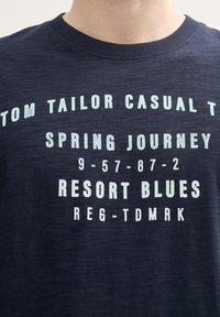 Jūras zila kokvilnas t-kreklā ar apaļu kakla izgriezumu, uz kura ir balti un gaiši zili uzraksti: "TOM TAILOR CASUAL T SPRING JOURNEY RESORT BLUES."