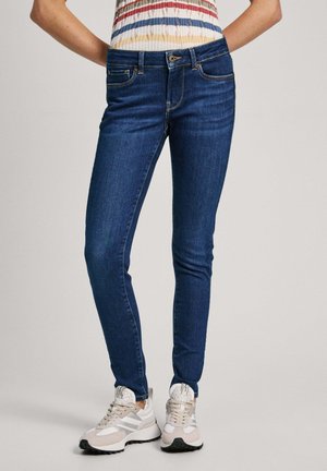 Personne portant un jean skinny bleu foncé, un haut à manches courtes rayé et des baskets blanches, debout devant un fond uni clair.