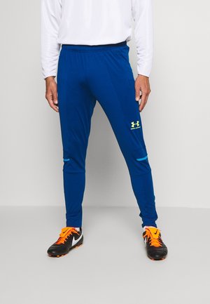 Mężczyzna ubrany w niebieskie sportowe spodnie Under Armour i czarne korki do piłki nożnej ze sznurówkami w kolorze pomarańczowym, stojący na szarym tle.