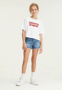 Balta saīsināta T-krekla ar sarkanu Levi’s logotipu, kopā ar gaiši ziliem džinsa šortiem. Modelis valkā baltas sporta kurpes un baltas potīšu zeķes.