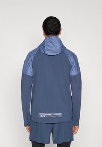 Αθλητικό μπουφάν Nike σε ναυτικό μπλε χρώμα με μακριά μανίκια, κουκούλα και λεπτομέρεια λογότυπου στο κάτω μέρος. Ομαλή υφή με αντίθετα πάνελ στους ώμους.