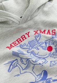 Grå hættetrøje med rød "MERRY XMAS" tekst og et blåt tegneserie-rensdyrdesign. Lavet af blødt stof med en struktureret overflade.