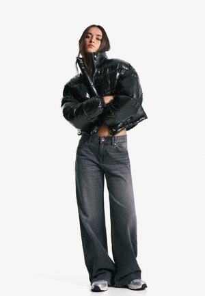 Zwarte glanzende cropped pufferjack met een hoge kraag, gecombineerd met losse donkergrijze jeans en grijze sneakers. Model staat met de armen over elkaar.