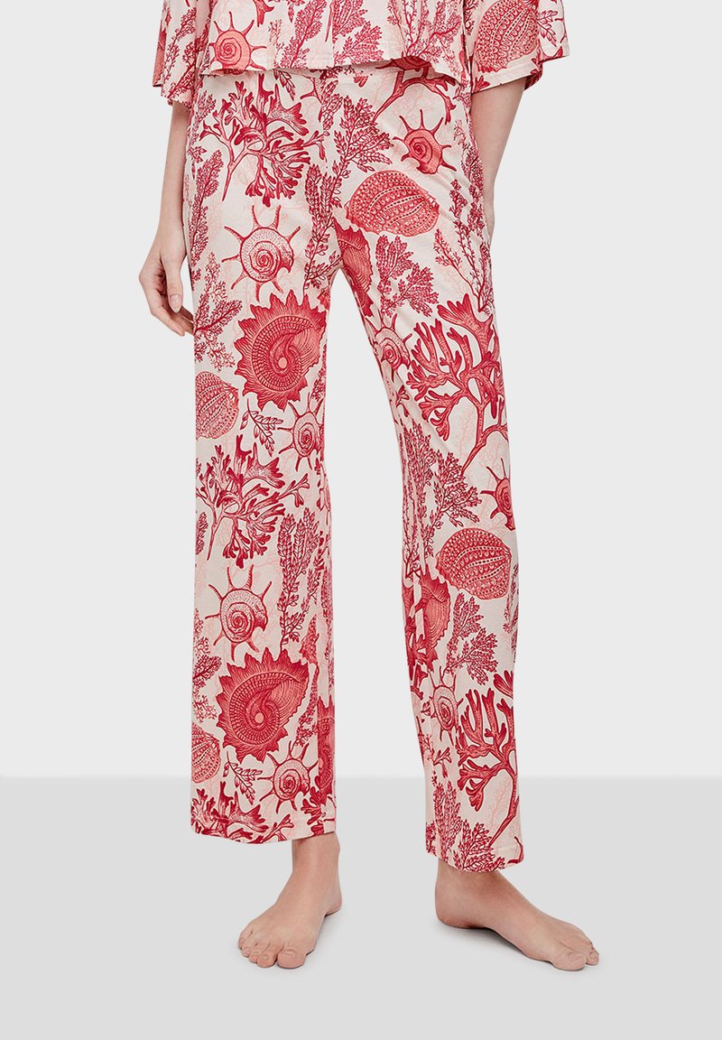 Femme portant un pantalon ample crème avec des motifs rouges de coquillages et de corail, pieds nus, debout devant un fond clair uni.
