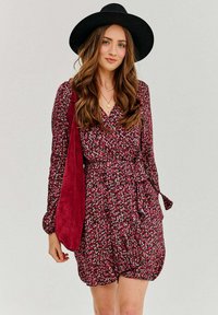 Kvetinové šaty v burgundskej farbe s malými bielymi a ružovými kvetmi, s dlhými rukávmi, viazanie v páse a jemnou textúrou; doplnené burgundskou kabelkou a čiernym klobúkom.