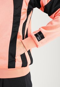 Růžová a černá sportovní mikina s dlouhými rukávy, s černými pruhy podél rukávů a štítkem na manžetě. Hladká textura.