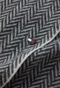 Tissu gris à motif en chevron avec une texture douce, présentant une petite épingle rectangulaire en noir, blanc et rouge positionnée sur le bord.