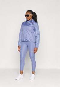 Lyse lilla vindjakke med ståkrave og strukturerede ærmer, parret med tætsiddende lyse lilla leggings og hvide sneakers. Sorte solbriller.