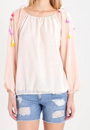 Kobieta ubrana w jasnoróżową, odkrywającą ramiona bluzkę z kolorowymi frędzlami na rękawach oraz jasnoniebieskie, postrzępione szorty dżinsowe.