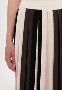 Gros plan d'une main à côté d'une jupe à plis verticaux avec des rayures noires, marron, beige et rose clair.