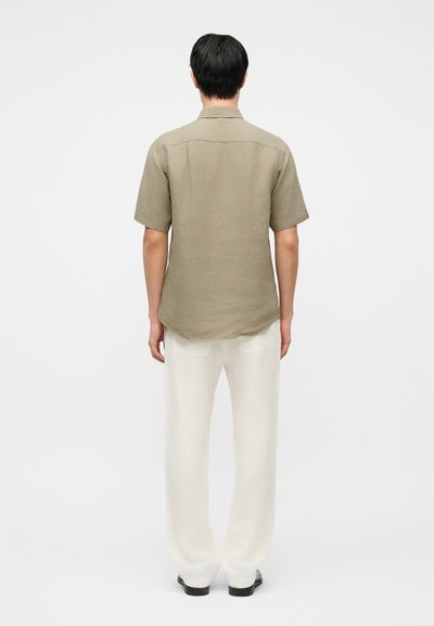 Άνδρας που στέκεται με την πλάτη στραμμένη, φορώντας κοντομάνικο μπεζ πουκάμισο, λευκό παντελόνι και μαύρα παπούτσια, σε απλό φωτεινό φόντο.