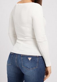 Haut à manches longues blanc à côtes associé à un jean bleu foncé avec un logo en forme de triangle sur la poche arrière. Texture lisse, design ajusté.