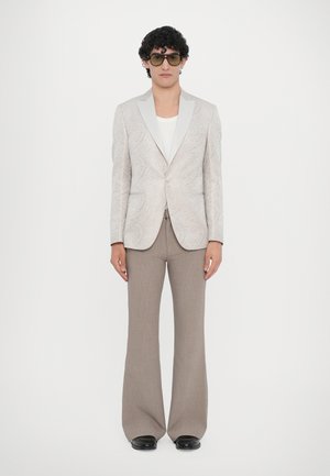 Light-colored paisley blazer with a contrasting white collar, paired with beige flared trousers. Model wears oversized, tinted sunglasses.