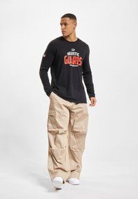 Zwarte longsleeve t-shirt met "HEVETIC GUARDS" in het rood bedrukt, gecombineerd met beige cargobroek. Model draagt witte sneakers.