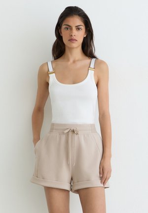 Kvinde med langt mørkt hår iført en hvid ærmeløs top med spænder og beige shorts med snører og opkrogede kanter, står op ad en hvid væg.