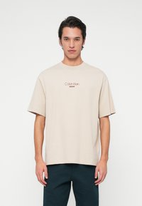 Vīriešu modelis, tērpies bēšā krāsā Calvin Klein Jeans T-krekl�ā un tumšās biksēs, stāv vērsts pretī kamerai uz vienkārša balta fona.