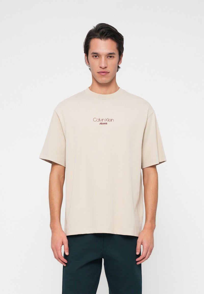 Vīriešu modelis, tērpies bēšā krāsā Calvin Klein Jeans T-kreklā un tumšās biksēs, stāv vērsts pretī kamerai uz vienkārša balta fona.