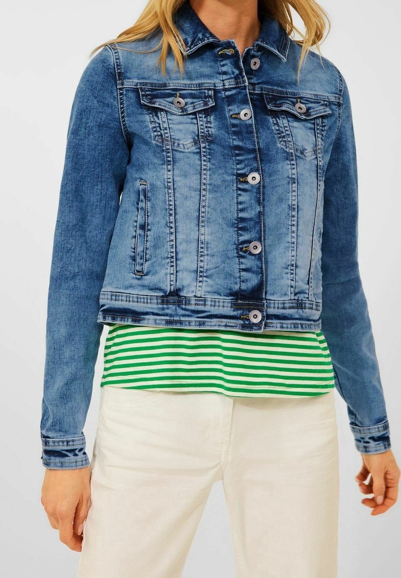 Woman wearing a blue denim jacket over a green and white striped shirt with off-white pants, standing against a plain light background.