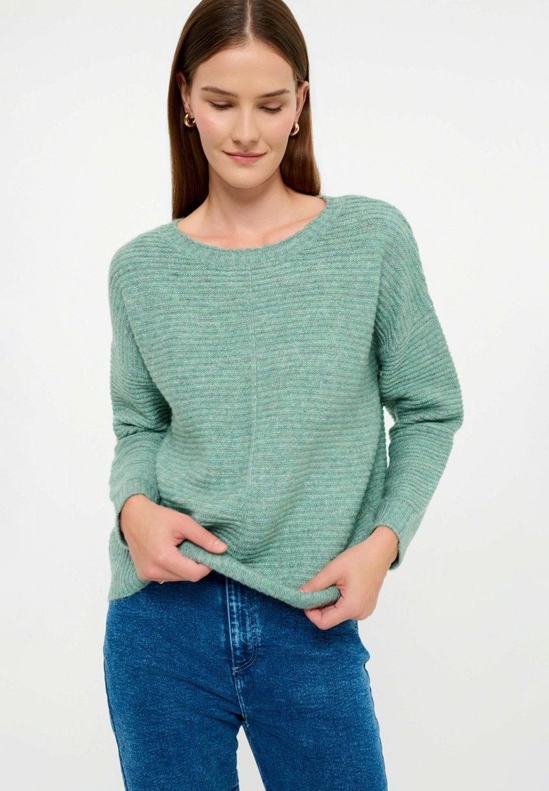 Maglione verde chiaro con un design strutturato a coste e un ampio scollo rotondo. Maniche lunghe e vestibilità rilassata abbinate a jeans in denim blu.