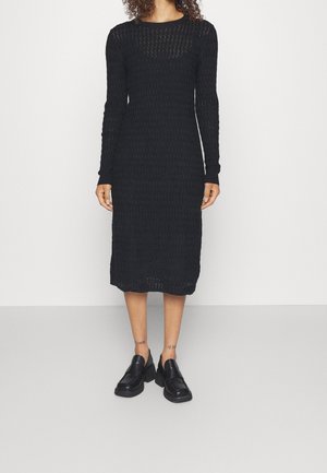 Femme portant une robe midi en maille texturée noire à manches longues et des mocassins noirs épais, debout sur un fond blanc uni.