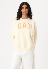 Kremfarget hettegenser med "GAP"-logo i en falmet oransje gradient. Har en avslappet passform, rund hals og ribbestrikkede mansjetter.
