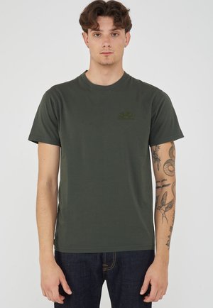 Jonge man met bruin haar, die een donkergroen T-shirt met korte mouwen en donkerblauwe jeans draagt, toont tatoeages op zijn rechterarm.