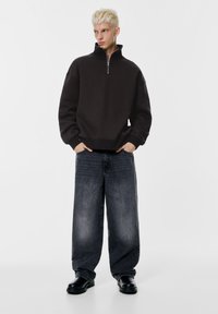 Mörkgrå, dragkedjeförsedd tröja med hög krage; kombinerad med lösa, randiga mörkblå jeans och svarta grova skor.