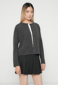 Jeune femme avec de longs cheveux ombrés portant un cardigan boutonné gris foncé sur un haut blanc et une jupe plissée noire, se tenant devant un fond uni.