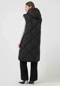 Svart quiltad ärmlös väst med huva, med diamantstickning och sidöppningar. Bärs över en vit långärmad topp och svarta byxor.