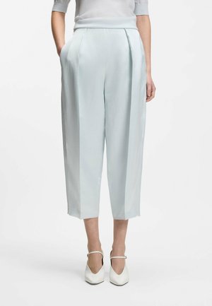 Femme portant un pantalon court taille haute bleu clair avec des plis, les mains dans les poches, associé à des escarpins blancs à bouts pointus avec des lanières à la cheville.