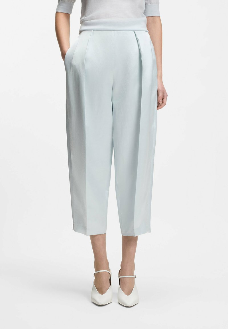 Femme portant un pantalon court taille haute bleu clair avec des plis, les mains dans les poches, associé à des escarpins blancs à bouts pointus avec des lanières à la cheville.