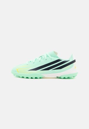 Chaussure de football vert clair avec des rayures noires, semelle blanche et petits crampons en dessous, vue de côté sur fond blanc.